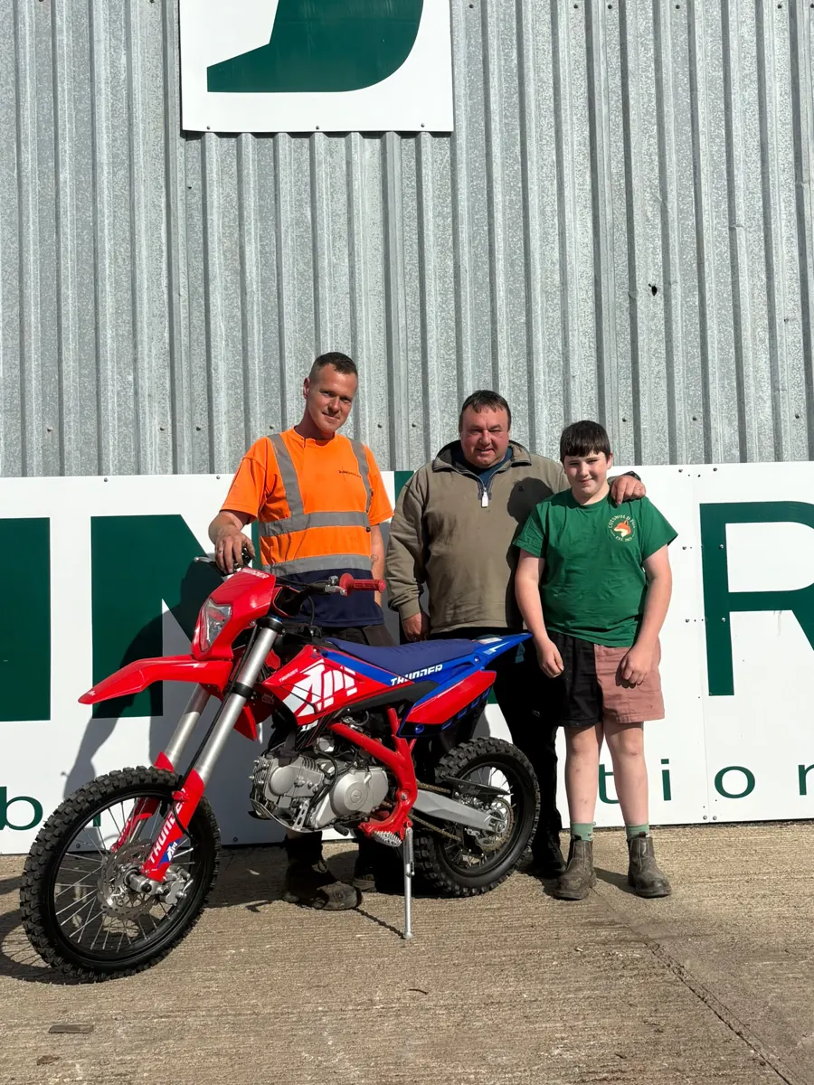 The winner’s son and two Dunn Bros team members with the new red and blue Apollo Thunder 125cc motocross bike in front of the Dunn Bros sign